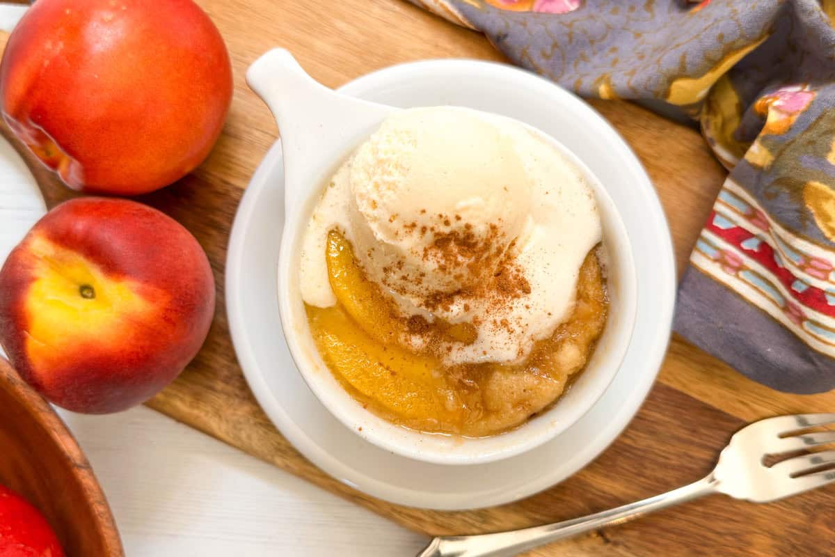 A bowl of peach cobbler topped with vanilla ice cream and a sprinkle of cinnamon sits on a wooden board, next to two fresh peaches, a fork, and a colorful cloth napkin—perfect inspiration for your next peach mug cake.