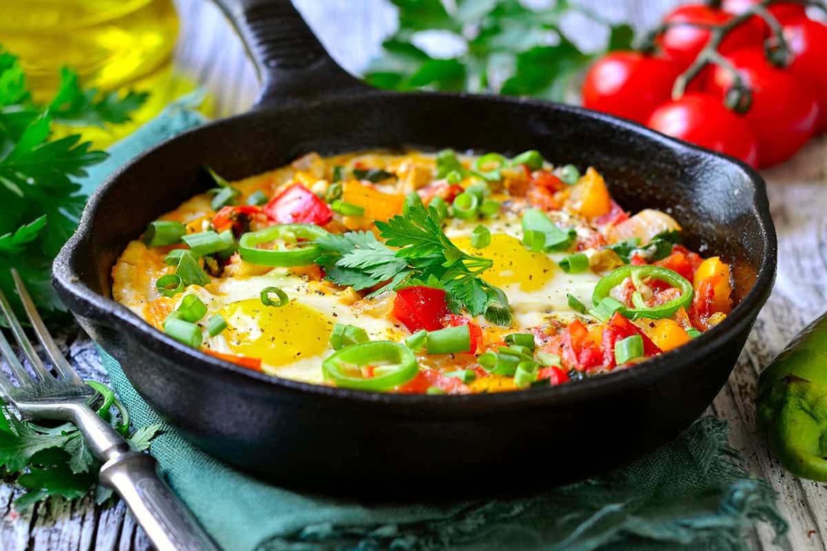 A skillet of shakshuka brimming with cooked eggs, tomatoes, peppers, and green onions, garnished with fresh herbs, rests on a rustic wooden table beside a fork and fresh vegetables.