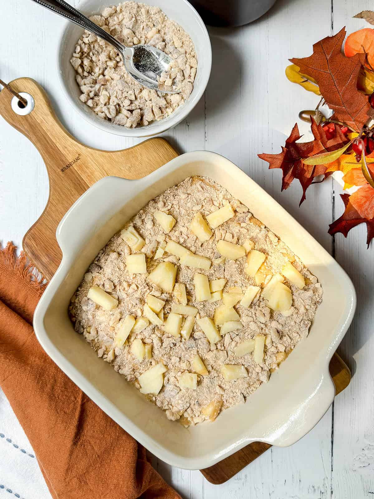 Unbaked caramel apple coffee cake layered with chopped apple and pecan streusel in a square baking pan.