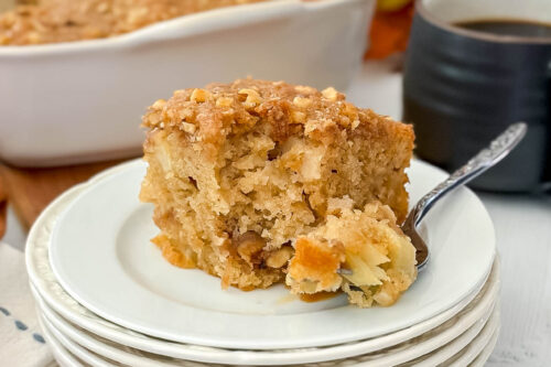 Slice of crumbly Apple Coffee Cake with visible nuts on a white plate, a mug of coffee, lit candle, and baking dish with the remaining cake in the background.