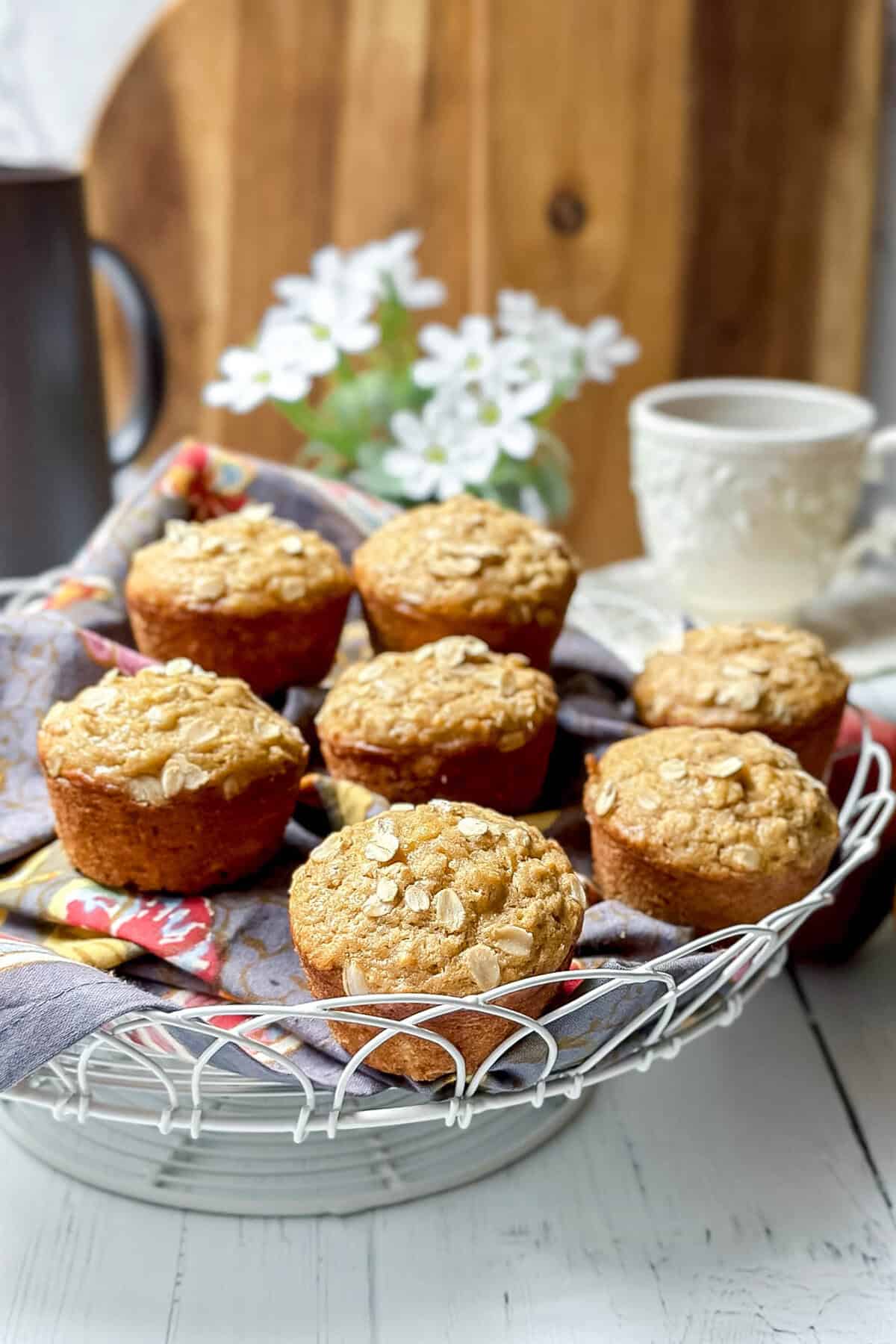 A wire basket holds seven apple oatmeal muffins topped with oats on a cloth napkin, with a white mug, black mug, and white flowers in the blurred background.
