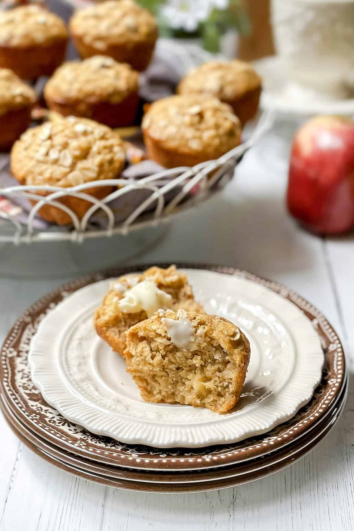 A muffin split in half with melting butter on a plate, with more apple oatmeal muffins in a basket and an apple in the background.
