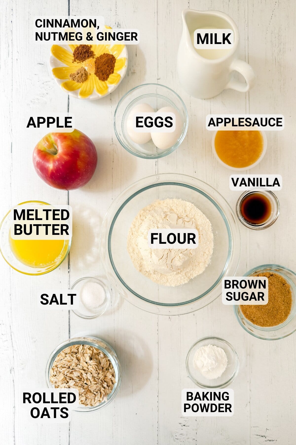 Various labeled baking ingredients for apple oatmeal muffins, including flour, eggs, apple, brown sugar, melted butter, rolled oats, baking powder, applesauce, vanilla, cinnamon, nutmeg, ginger, milk, and salt on a white surface.