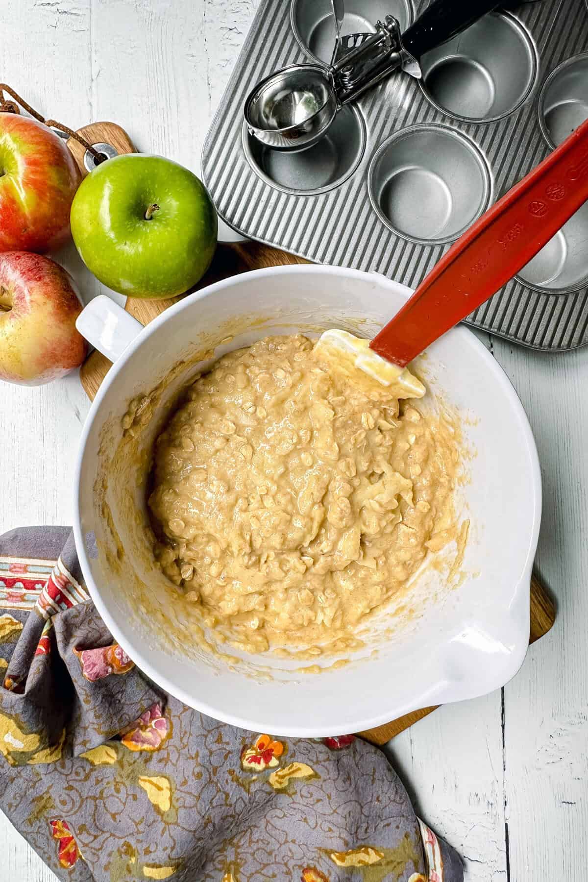 A mixing bowl with apple oatmeal muffins batter, a red spatula, apples, a muffin tin, an ice cream scoop, and a patterned cloth on a white surface.