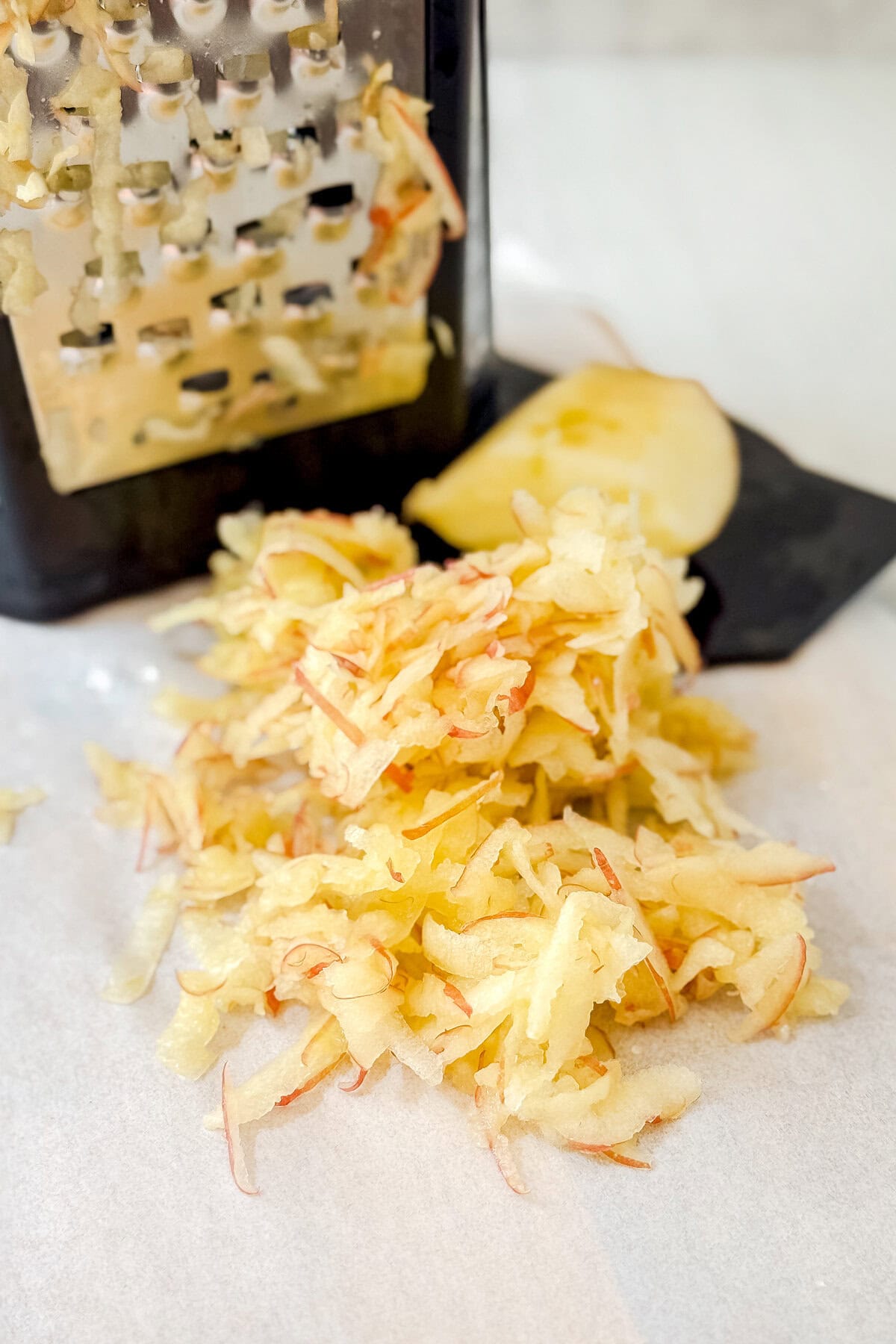 A pile of freshly grated apple sits on parchment paper in front of a box grater and a sliced apple, ready to be mixed into delicious apple oatmeal muffins.