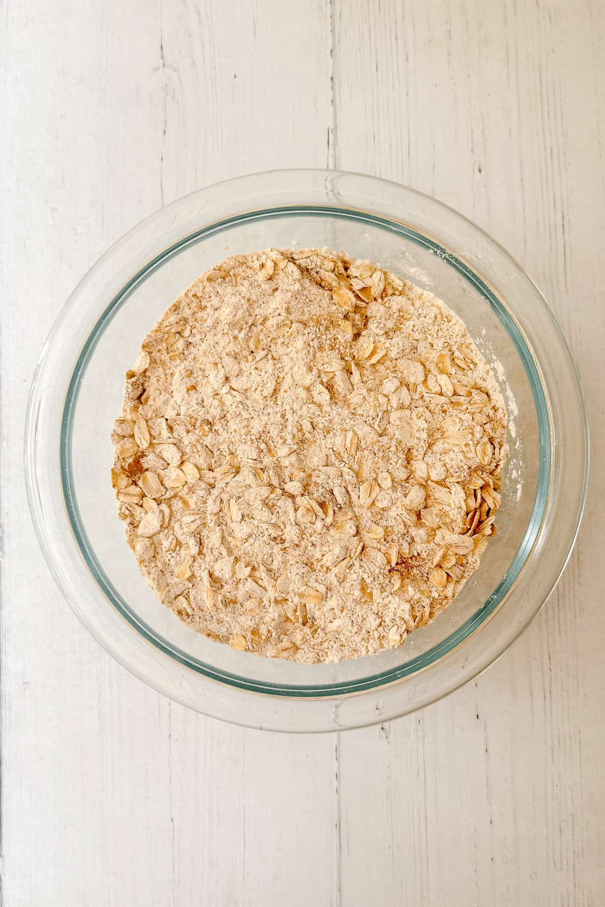 A glass bowl containing a dry mixture of oats and flour for apple oatmeal muffins sits on a white wooden surface.