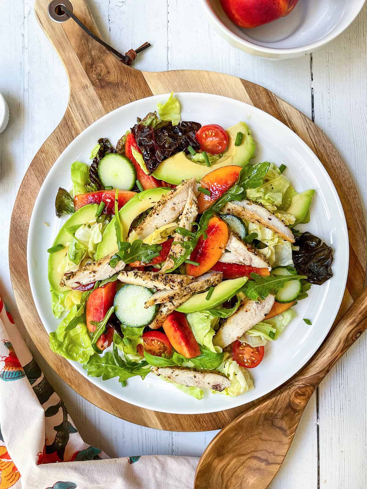 A plate of chicken salad with mixed greens, grilled chicken strips, cucumber, cherry tomatoes, avocado, and peach slices is served on a wooden board.