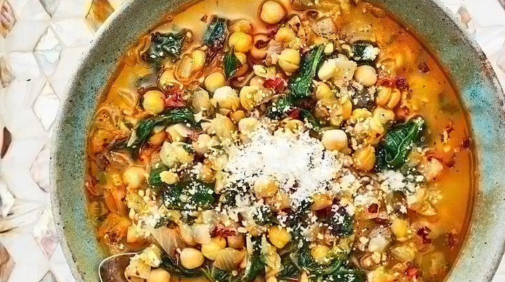 A bowl of chickpea and greens soup topped with grated cheese, alongside a spoon, sits on a patterned surface next to a glass of amber liquid—an inviting scene inspired by Mediterranean Diet Skillet Dinners.