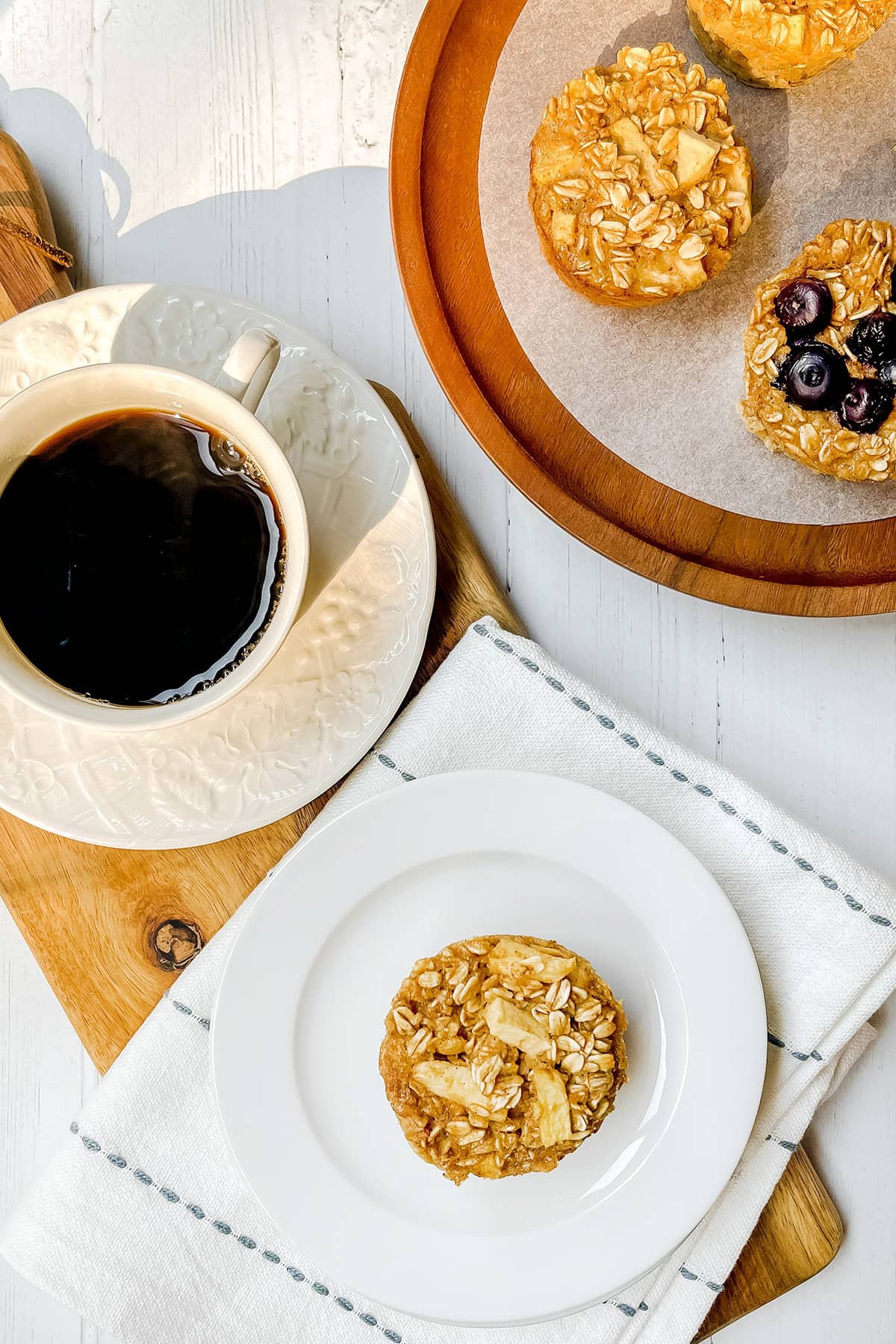 Top view of an apple baked oatmeal cup on a white with a cup of coffee nearby and a platter of plain and blueberries baked oatmeal cups.