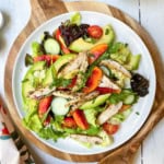 A white plate of chicken salad with grilled chicken, lettuce, cucumbers, avocado, cherry tomatoes, peach slices, and leafy greens sits on a wooden board.