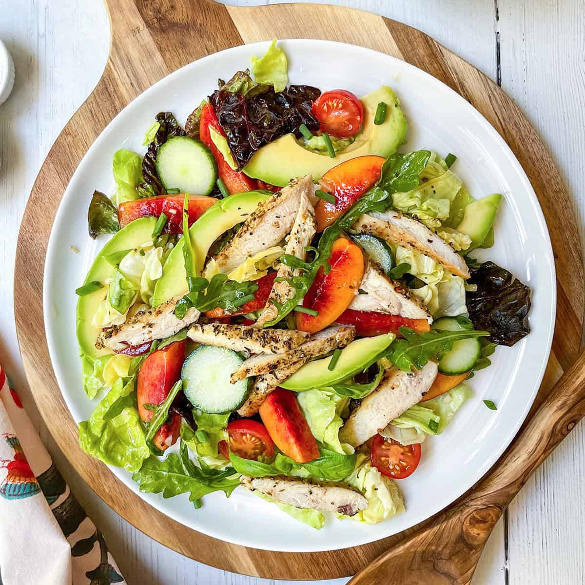 A white plate of chicken salad with grilled chicken, lettuce, cucumbers, avocado, cherry tomatoes, peach slices, and leafy greens sits on a wooden board.