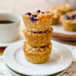 Square image of three stacked baked oatmeal cups on a plate with a cup of coffee in the background.