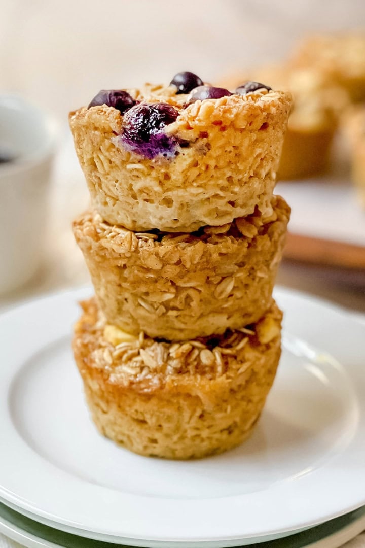 Square image of three stacked baked oatmeal cups on a plate with a cup of coffee in the background.