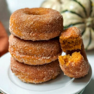 Cinnamon sugar baked pumpkin donuts stacked on plate with another on its side with a bite missing.