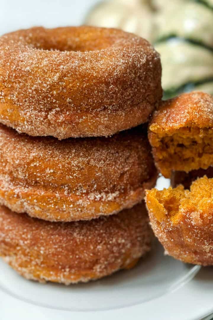 Cinnamon sugar baked pumpkin donuts stacked on plate with another on its side with a bite missing.