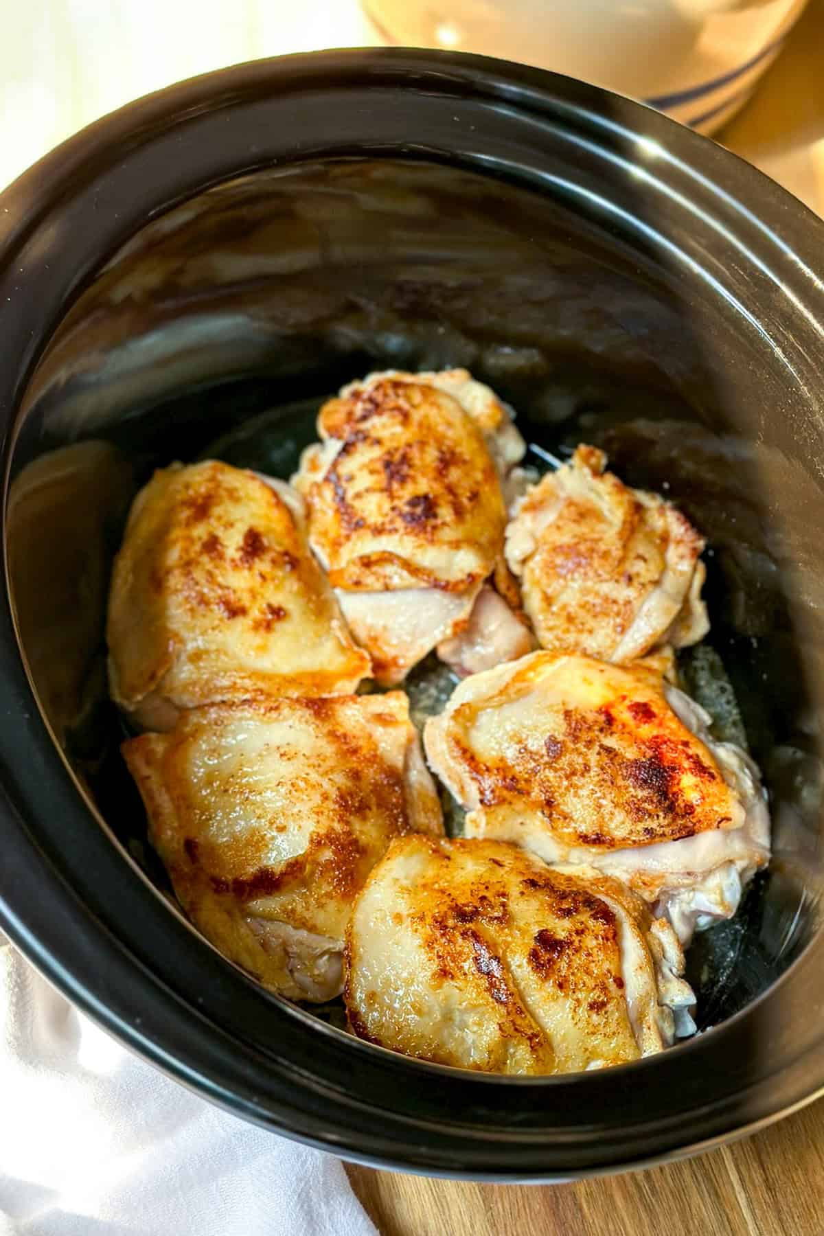 Six browned slow cooker chicken thighs inside a black slow cooker, shown on a wooden surface with a white cloth partially visible.