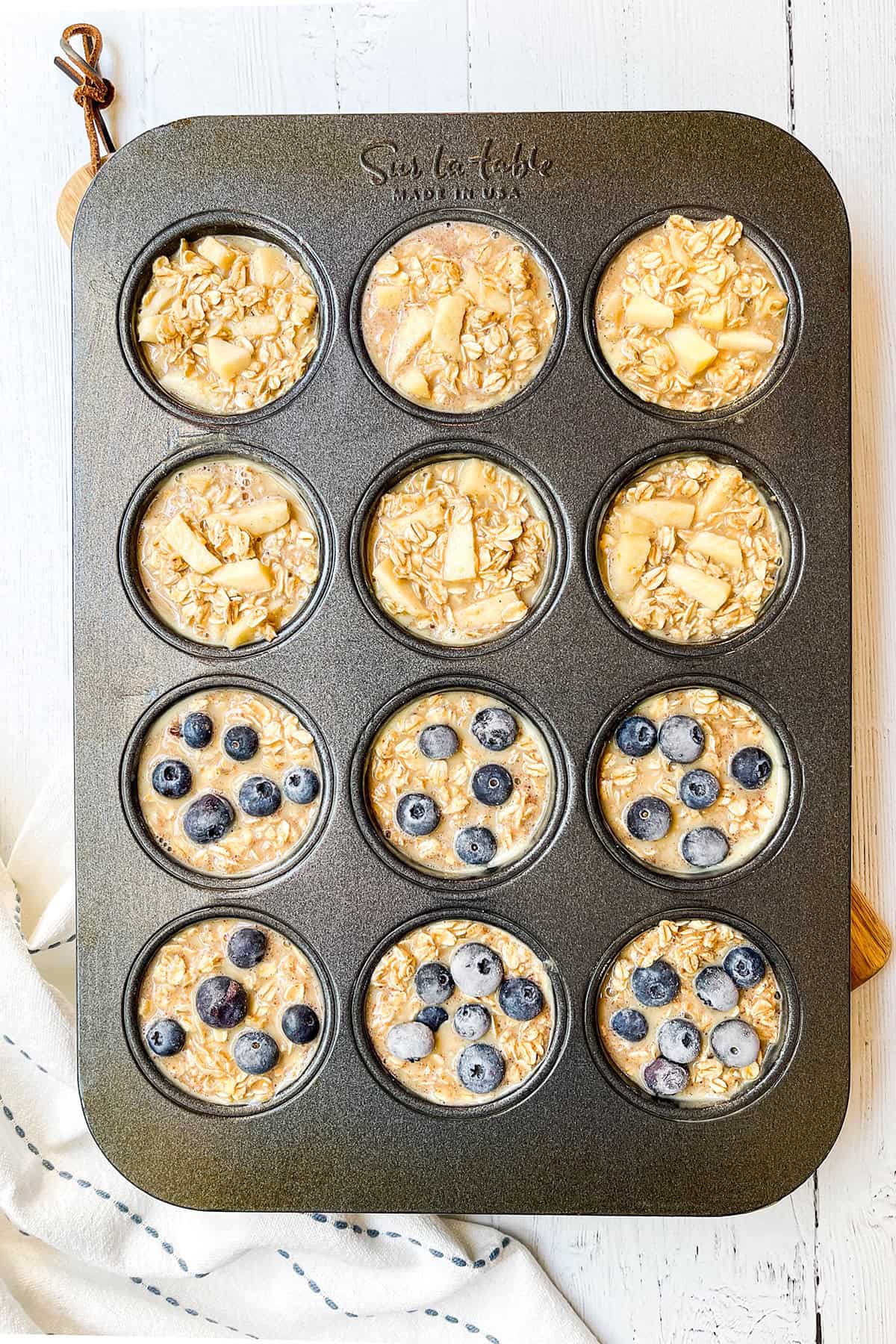Unbaked oatmeal cups in a muffin tin, filled with oats, apples, and blueberries before baking.