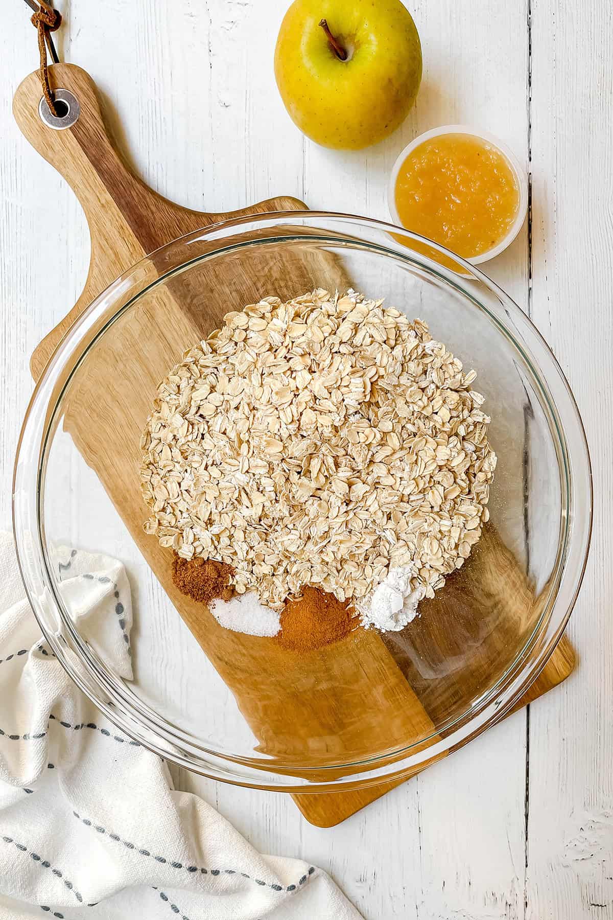 Glass mixing bowl with rolled oats, cinnamon, baking powder, and salt, ready to make baked oatmeal cups. A yellow apple and applesauce sit nearby.