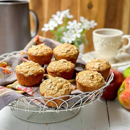 A wire basket with seven apple oatmeal muffins topped with oats sits on a cloth napkin, with a ceramic cup, pitcher, white flowers, and apples in the background on a white table.