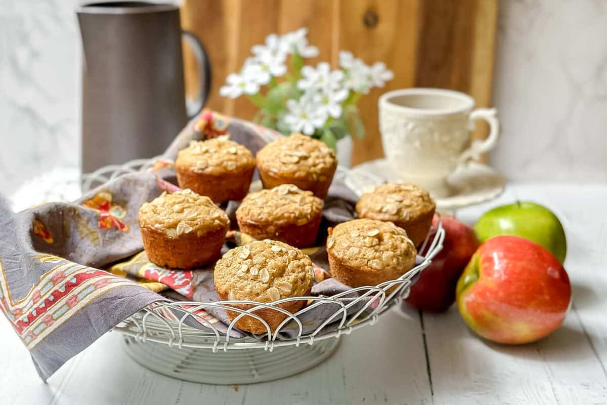 A wire basket with seven apple oatmeal muffins topped with oats sits on a cloth napkin, with a ceramic cup, pitcher, white flowers, and apples in the background on a white table.