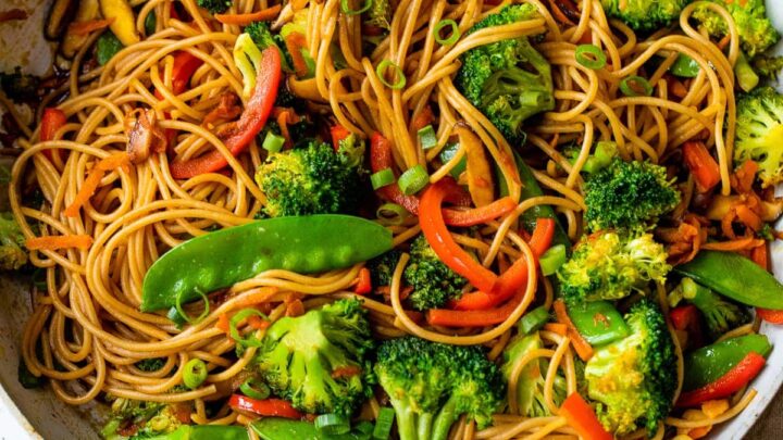 A skillet filled with vegetable lo mein—noodles, broccoli, snap peas, red bell peppers, and carrots—served with sliced green onions and sauce on the side. One of our favorite 20-Minute Fall Dinners for a quick, flavorful meal.