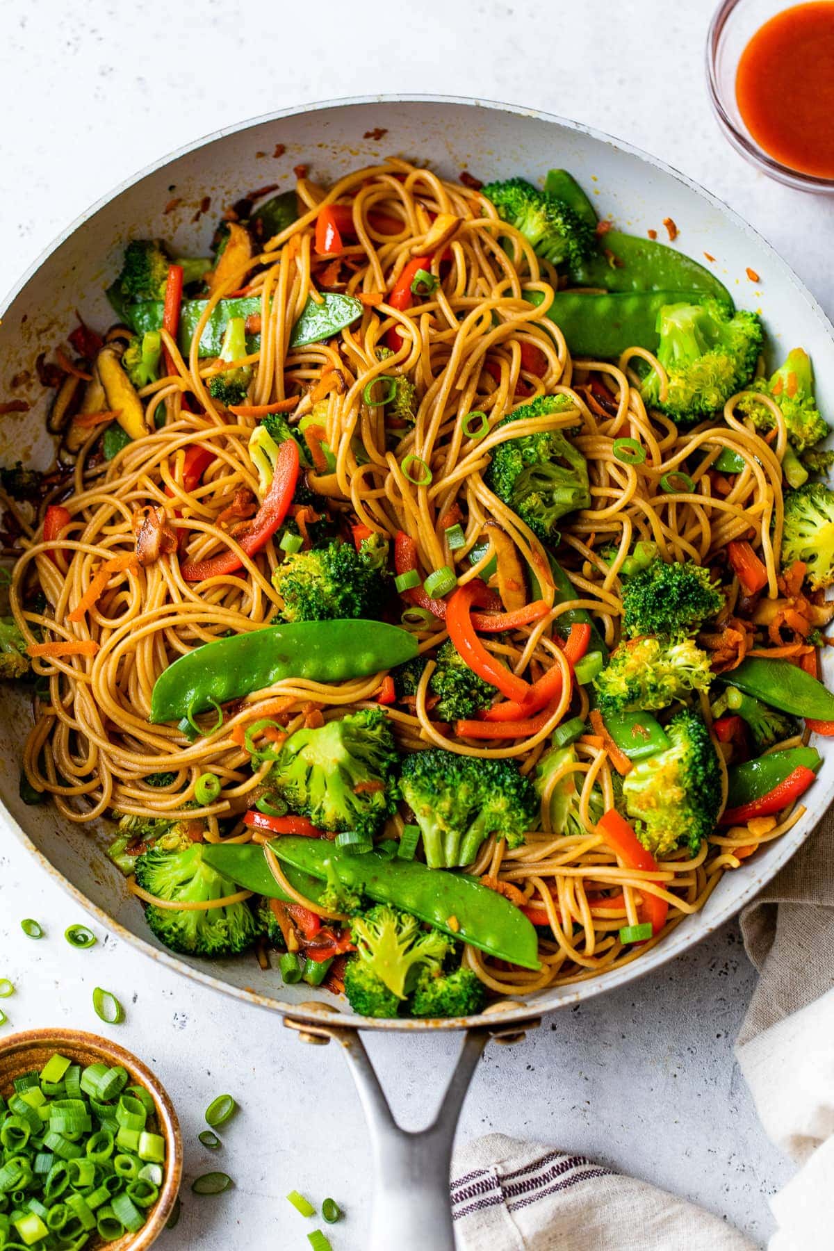 A skillet filled with vegetable lo mein-noodles, broccoli, snap peas, red bell peppers, and carrots-served with sliced green onions and sauce on the side. One of our favorite 20-Minute Fall Dinners for a quick, flavorful meal.