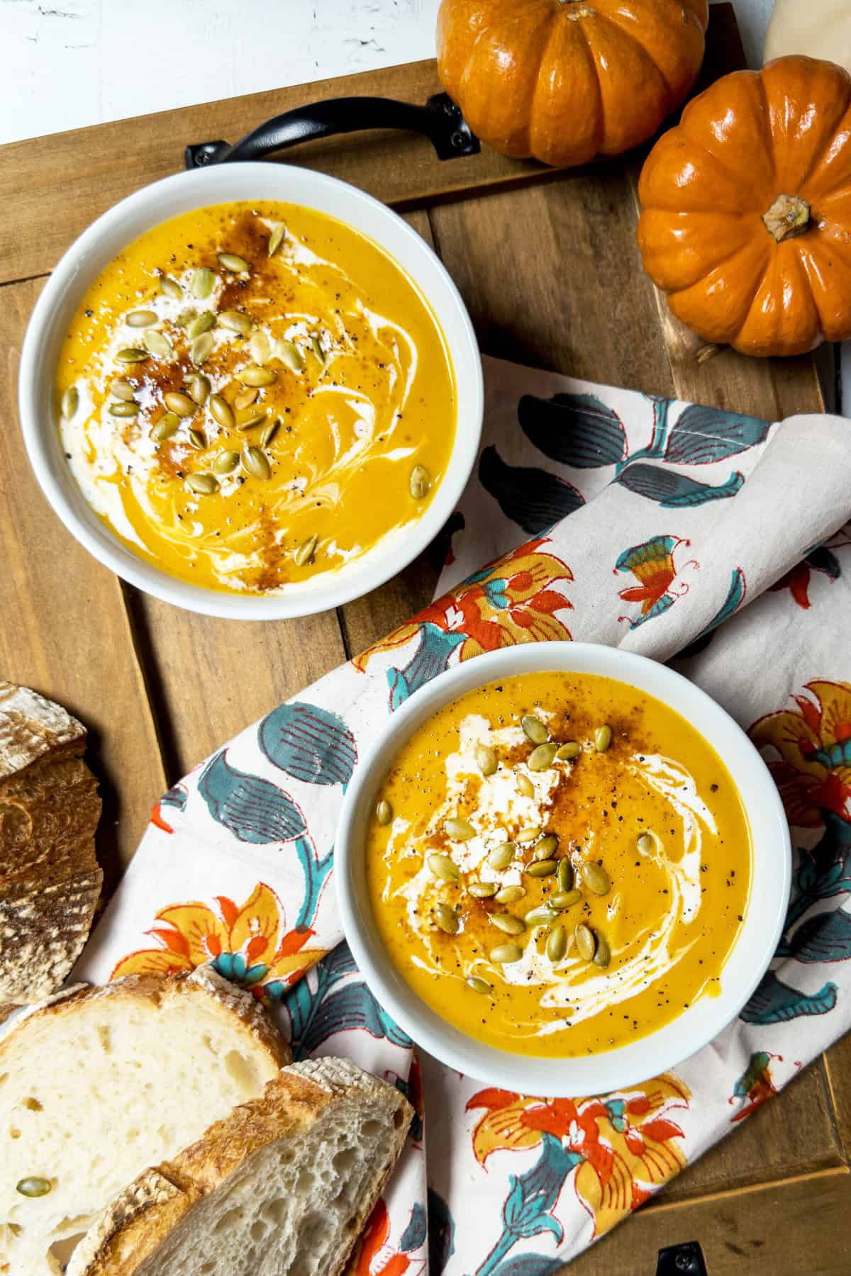Two bowls of pumpkin soup, inspired by the Panera autumn squash soup recipe, topped with cream, seeds, and seasoning sit on a floral napkin with slices of bread and decorative pumpkins nearby.