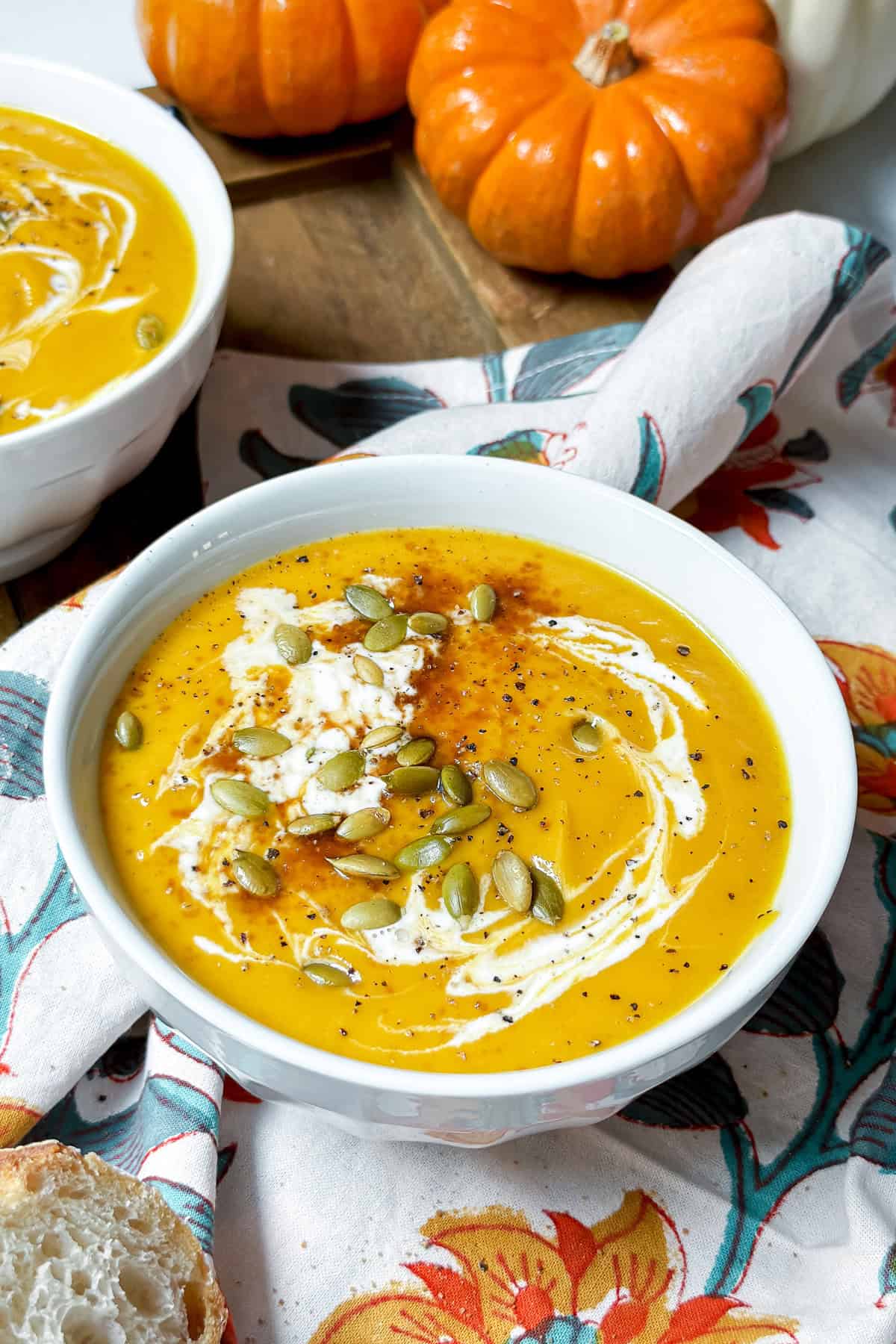 A bowl of creamy orange soup inspired by the Panera Autumn Squash Soup recipe, topped with pepitas, black pepper, and a swirl of cream, set on a floral napkin with pumpkins in the background.