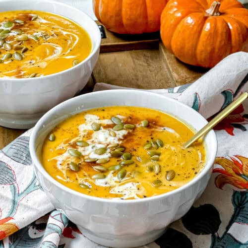 Two bowls of pumpkin soup inspired by the Panera autumn squash soup recipe, topped with seeds and cream, set on a floral napkin, with small pumpkins and a plate of pumpkin seeds in the background.