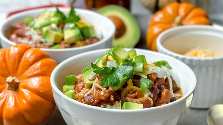 Bowl of pumpkin turkey chili topped with avocado, cheese, and herbs, served with pumpkins and fall décor.