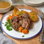Slow Cooker roast beef, potatoes, and carrots on a bue and white plate with gravy in a bowl nearby.