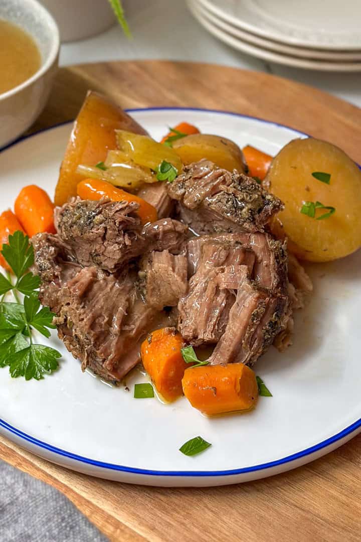 Slow Cooker roast beef, potatoes, and carrots on a bue and white plate with gravy in a bowl nearby.