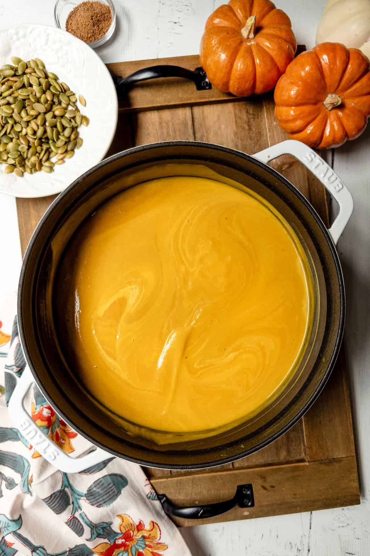 A pot of creamy pumpkin soup, inspired by the Panera autumn squash soup recipe, sits on a wooden board, surrounded by small pumpkins, a bowl of pumpkin seeds, and a small dish of spices.