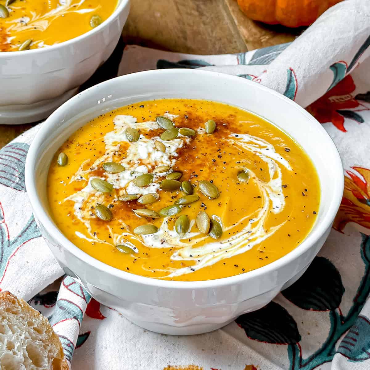 A white bowl filled with creamy orange soup, inspired by the Panera autumn squash soup recipe, is topped with a swirl of cream, pumpkin seeds, and black pepper, and sits on a floral napkin next to a slice of bread.