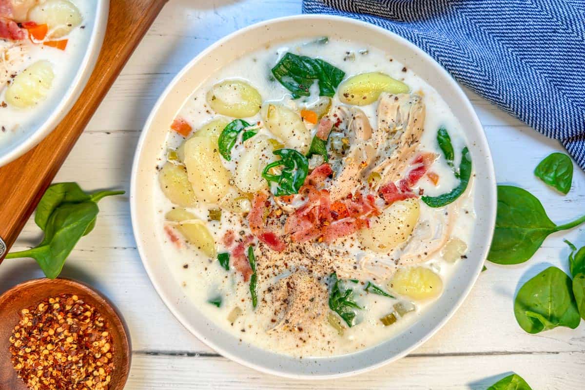 A creamy bowl of chicken gnocchi soup with spinach, shredded chicken, and crispy bacon bits. Topped with black pepper, it's served on a white wooden table adorned with scattered spinach leaves and a small bowl of red pepper flakes nearby. A blue cloth napkin lies to the side.