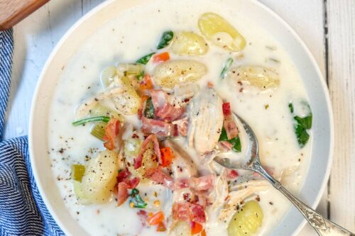 A bowl of creamy chicken gnocchi soup brimming with shredded chicken, spinach, and diced tomatoes. A spoon rests in the bowl. A wooden tray with red pepper flakes is nearby, and a blue cloth graces the white table adorned with scattered leafy greens.