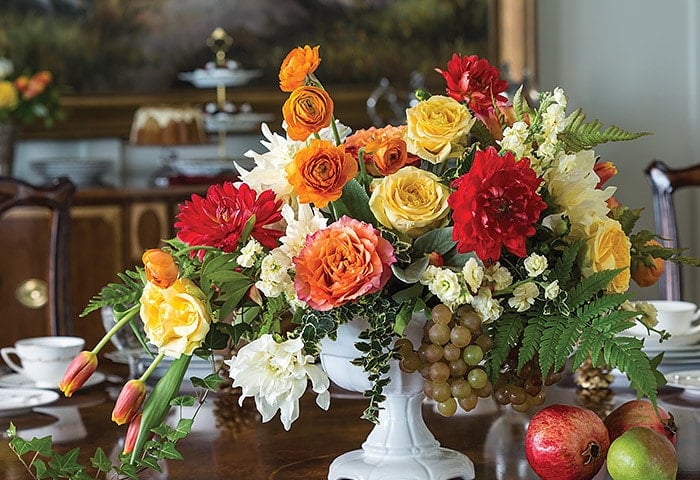A floral arrangement with red, yellow, orange, and white flowers, green leaves, grapes, and pomegranates sits on a wooden table set for a cozy November Afternoon Tea.