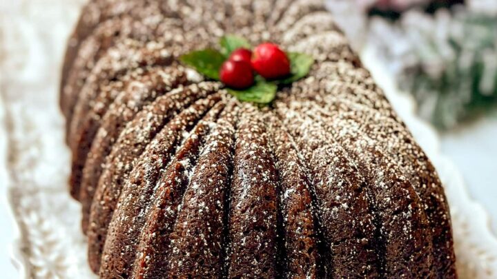 Closeup side view of unsliced Gingerbread Loaf Cake on a white lace serving dish with powdered sugar, cranberries, and mint leaves.