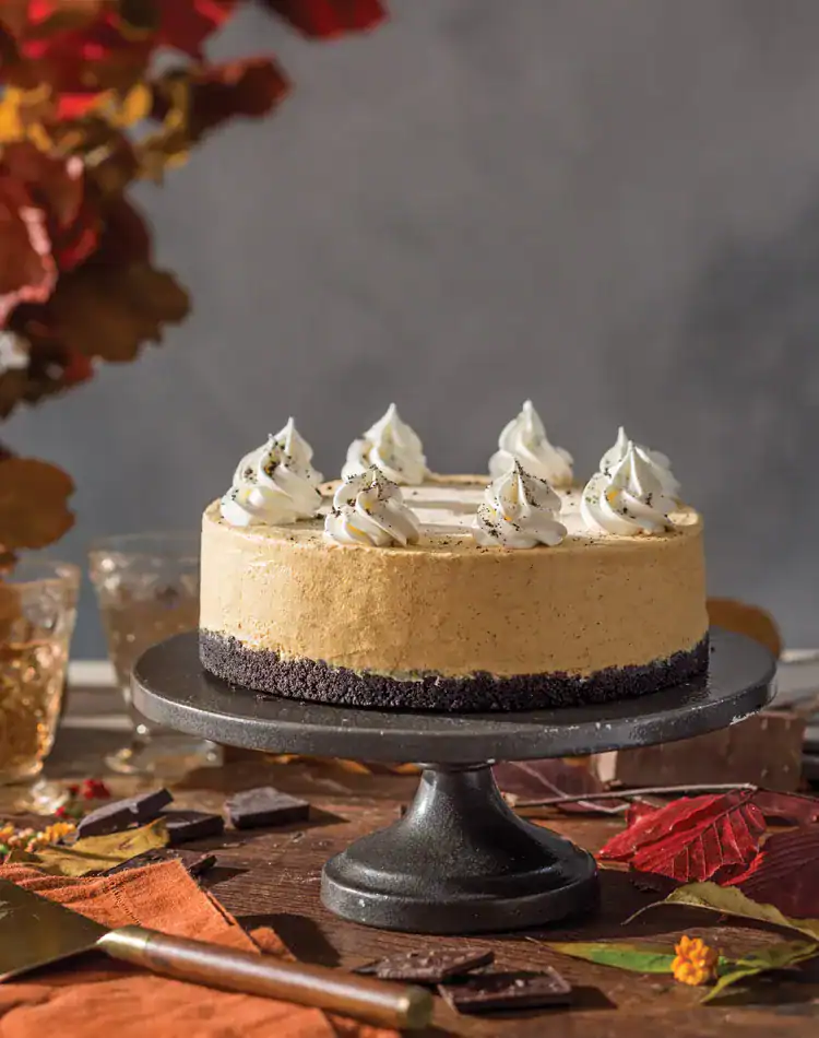 A pumpkin cheesecake with a chocolate crust and whipped cream topping sits on a black cake stand, perfect for a cozy November Afternoon Tea, surrounded by autumn leaves and elegant table settings.