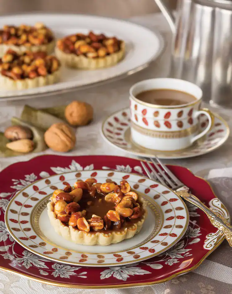 A nut tart sits on a decorative plate with a fork beside it; in the background are more tarts, a cup of coffee, metal pot, and whole walnuts-perfect for a cozy November Afternoon Tea on a patterned tablecloth.