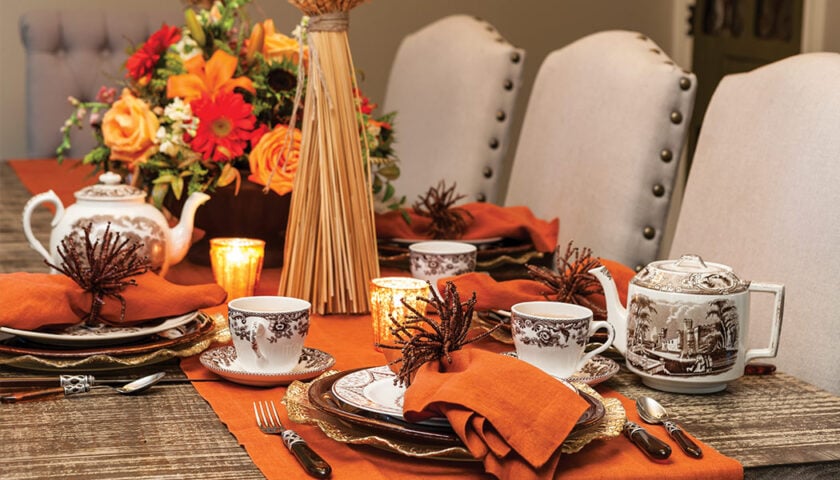 A dining table set with orange napkins, patterned china, teapots, floral centerpiece, and lit candles creates a cozy November Afternoon Tea with an autumn-themed arrangement.