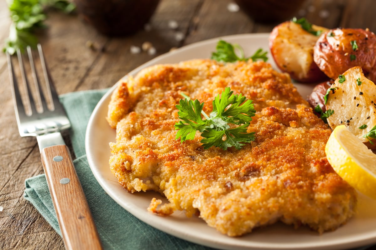 Chicken schnitzel on a plate with parsley, herbed potatoes and a lemon wedge.