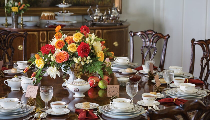 A formal dining table set for a November Afternoon Tea with china, glasses, red napkins, soup bowls, and a vibrant centerpiece of yellow, orange, and red flowers. In the background sits a sideboard featuring tempting desserts and fresh fruit.