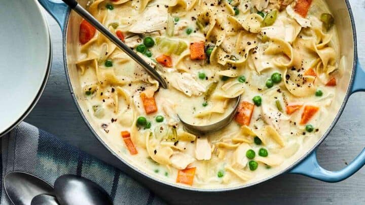 Top view of creamy chicken noodle soup in a light blue Dutch oven with serving ladel over a white wood background.