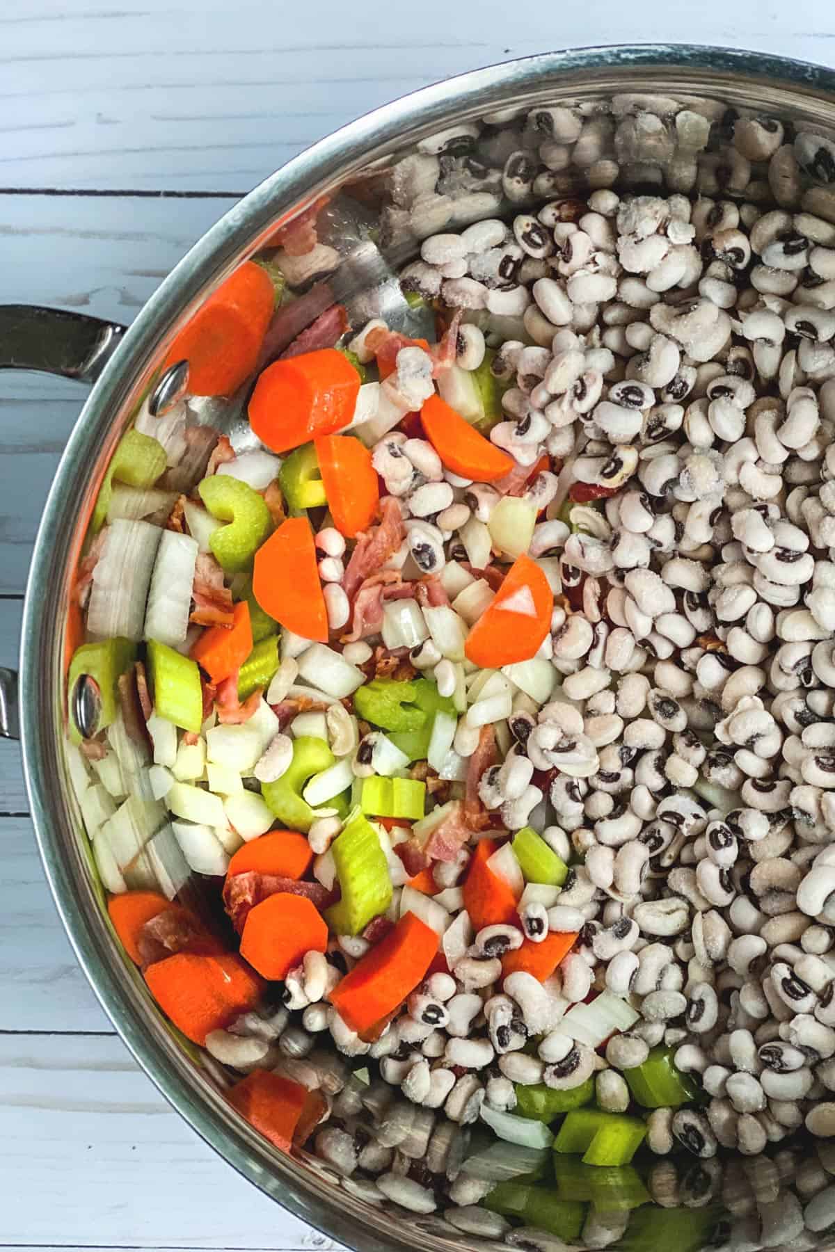 A stainless steel pot filled with chopped celery, carrots, onions, and black-eyed peas sits ready for a festive New Year's Day Black Eyed Peas Soup. The vegetables are neatly arranged on the left, while the peas fill the right side of the pot against a rustic wooden background.