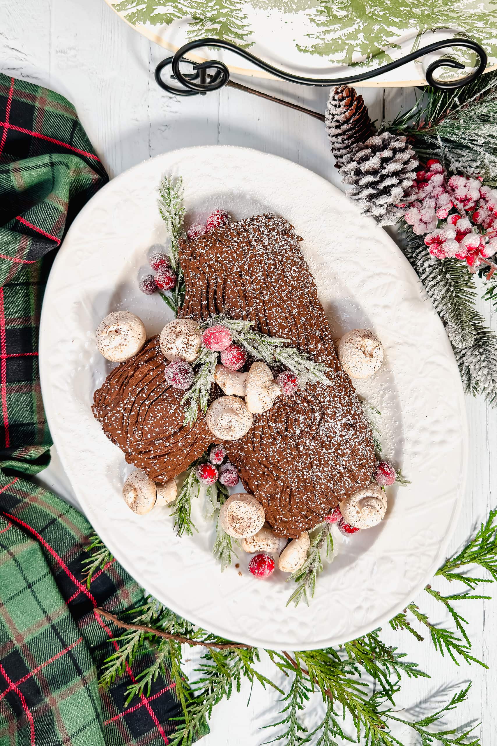 Top view of Yule Log cake with meringue mushrooms and sugared cranberries and rosemary on a white oval platter.