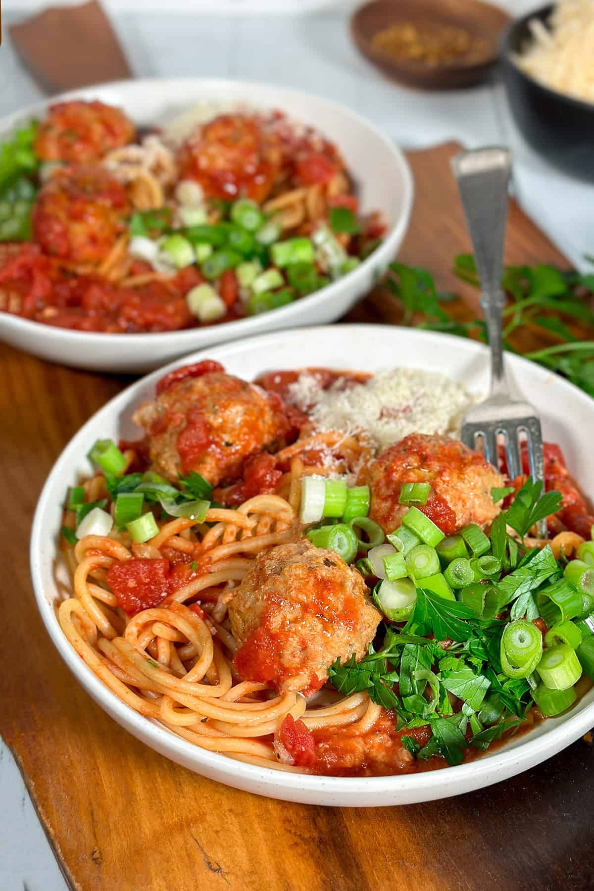 Two bowls of crockpot turkey meatballs and spaghetti on a wooden serving board and garnished with chopped parsley, Parmesan, and green onions.