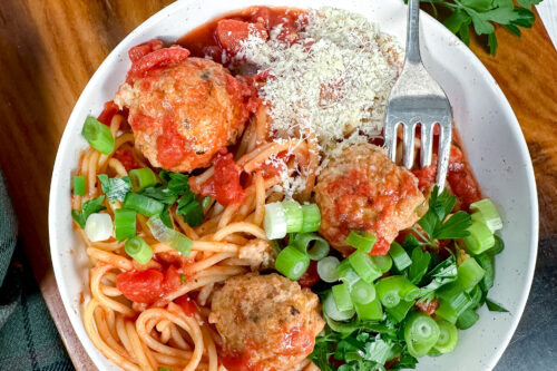 A bowl of spaghetti with Crockpot Turkey Meatballs, tomato sauce, grated cheese, chopped green onions, and parsley, with a fork resting in the bowl.
