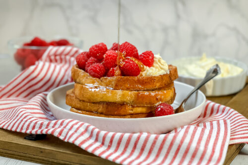 Horizontal side view of baked French Toast, stacked with drizzled honey, fresh raspberries, and lemon mascarpone.