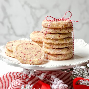 Square image of stacked Christmas slice and bake cookies with red sprinkles on a dessert stand tied with a red and white ribbon.